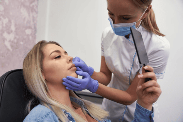 Woman holding a mirror while an aesthetic practitioner examines her facial features during a medical spa consultation.